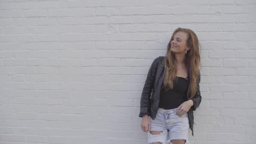 Woman leans against white brick wall smiles and plays with hair