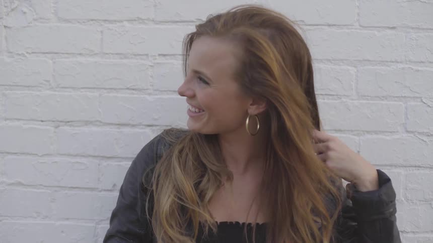 A female stands in front of a white brick wall laughing and fixing her highlighted hair as it gets blown by the wind