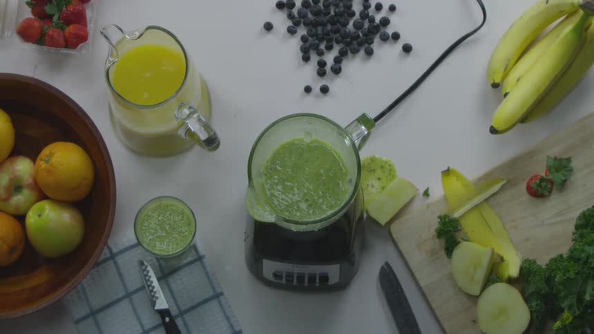 A person picks up a glass from a recently blended smoothie which sits on a prep table full of fruit