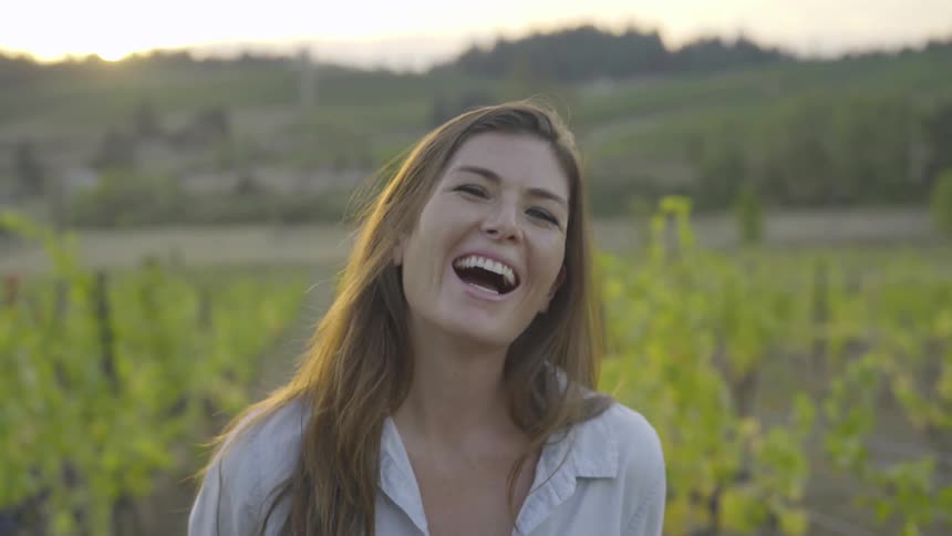 A woman stands in a vineyard with young vines behind her laughing and smiling