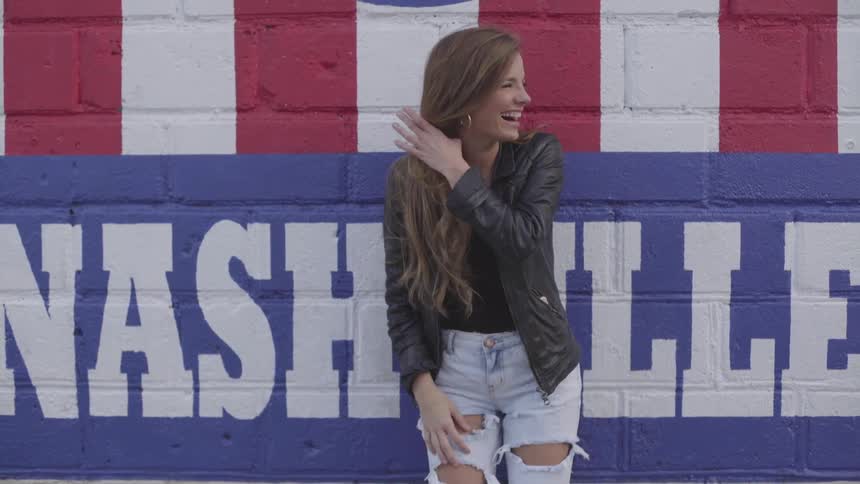 A young woman wearing torn jeans leans against a painted wall laughing and playing with her long hair