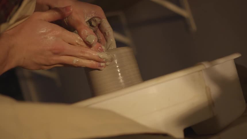 A potter leans forward over a pottery wheel and uses his hands and fingers to shape the wet clay