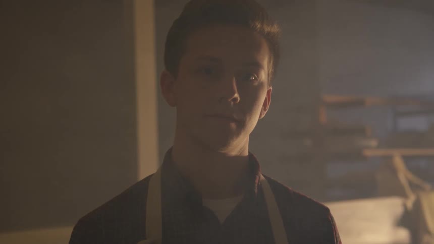 A young white man wearing an apron standing in a dimly lit workshop