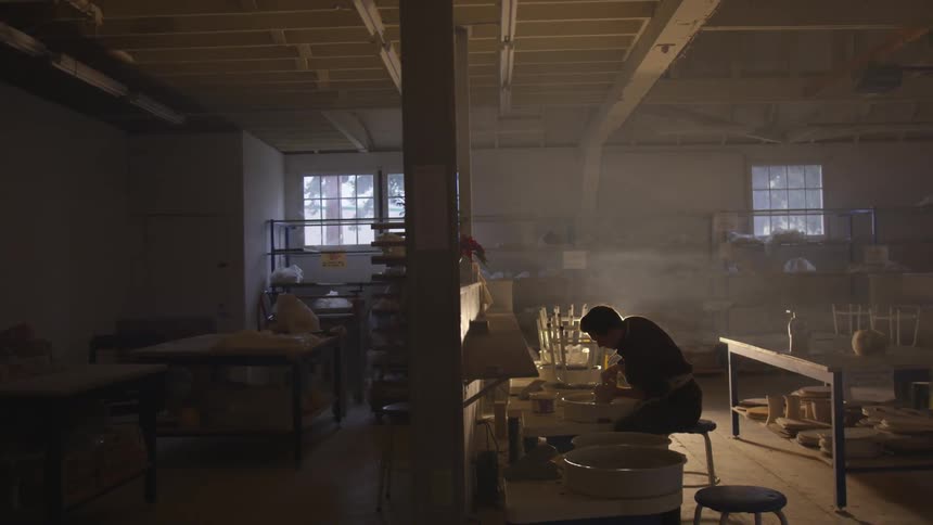 A sculptor sits and works at a table in a dark warehouse