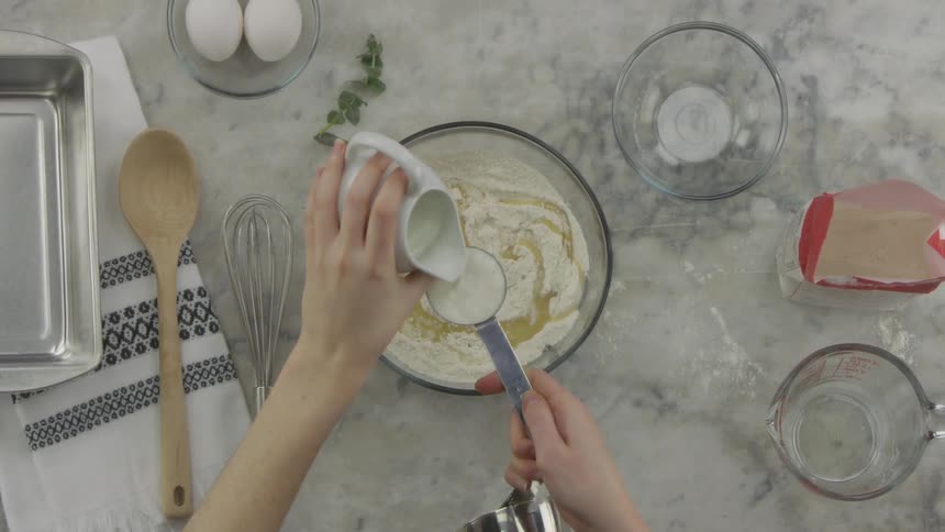 A person is measuring out milk from a white glass container into a stainless steel measuring cup and then adding it to an egg and flour mixture in a glass bowl