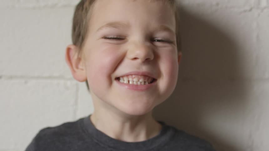 A young boy standing against a white wall smiles big and closes his eyes