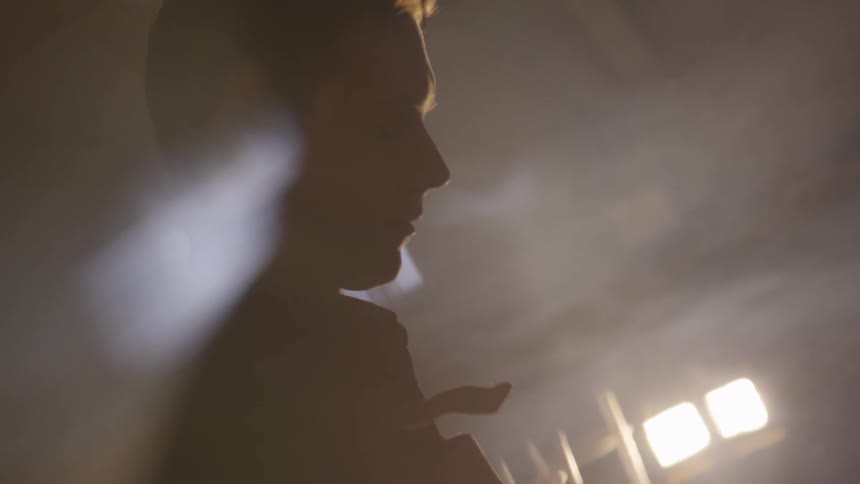 A young man stands in a dark smoky room with his eyes closed and his head looking down