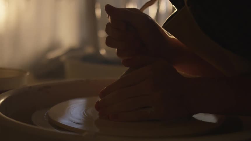 A man leans over a pottery wheel as he uses his hands to wet and shape clay