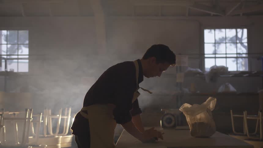 A man in an apron works on a material on a table in dark warehouse
