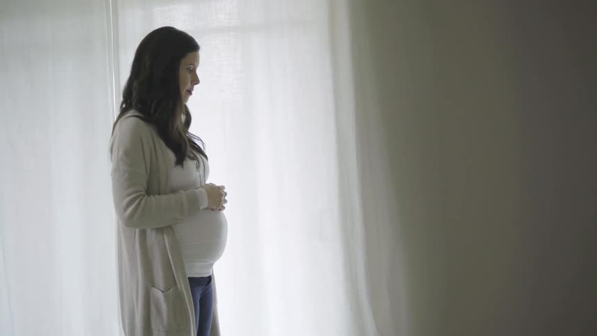 A pregnant woman stands still with her hands atop her protruding belly as she appears to be lost in thought