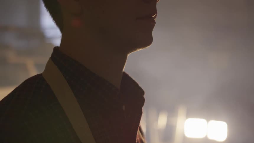 A person in a collared shirt stands still adjacent to some bright lights and tall windows