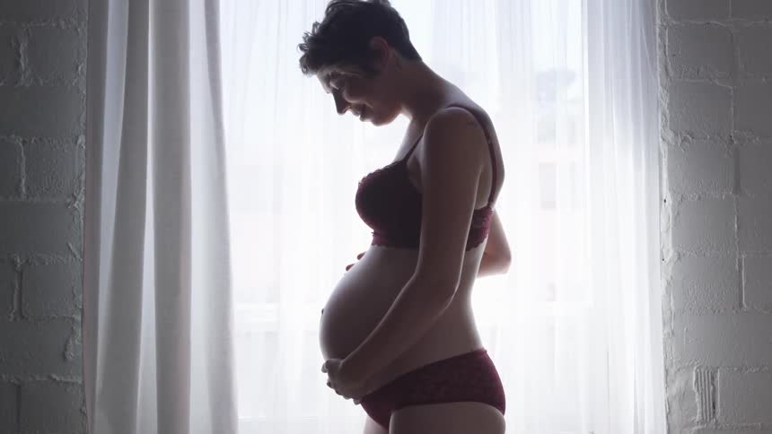 A pregnant woman poses in front of a window in her lingerie looking down and caressing her belly