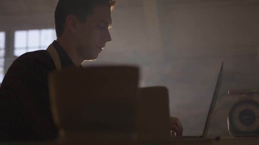 A man wearing an apron sits at a table typing on a laptop