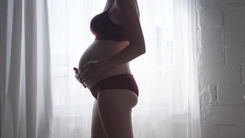 Pregnant woman in her underwear standing near window rubbing her belly