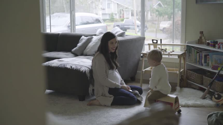 A young woman who is pregnant kneels on the floor as she watches and encourages a young toddler who is scooting along the floor of their living room on a wheeled toy