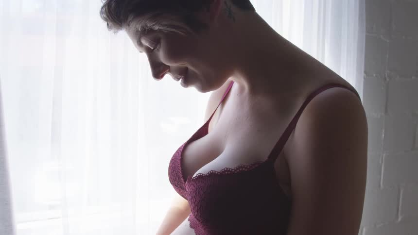 A woman in a black bra stands still in front of a window as she looks downward and subtly smiles
