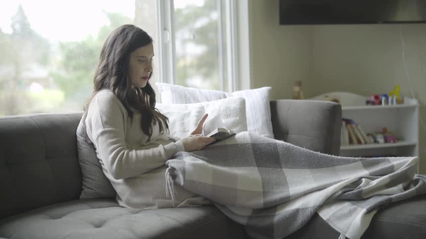 A woman reads on a sofa with a blanket on her legs