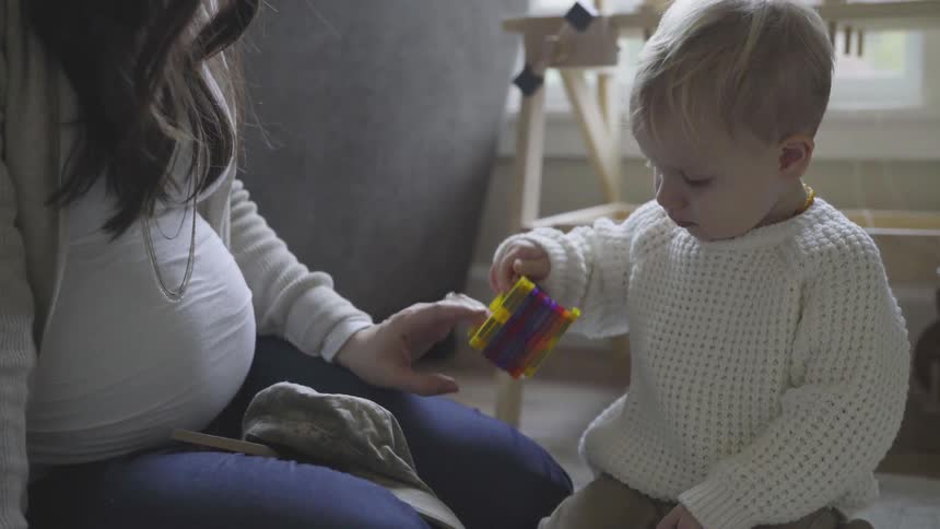 A small toddler in a knit sweater sits on the ground an plays with some small magnetic tile toys while a pregnant woman hands the child some more