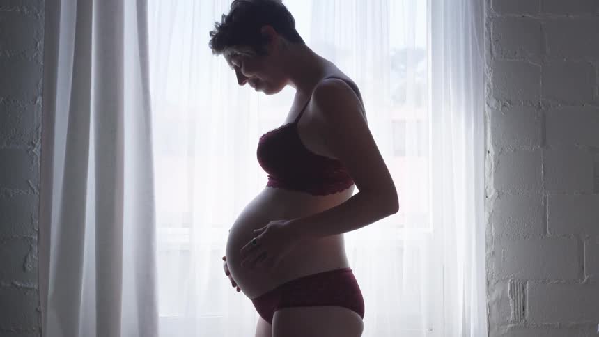 A pregnant woman dressed in a bra and underwear rubs her belly bump in front of a window