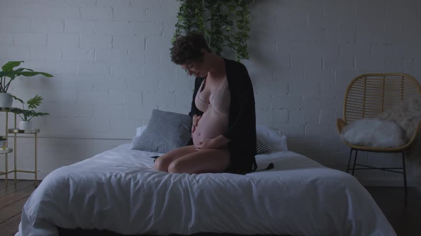 Woman sits on a bed holding her pregnant stomach