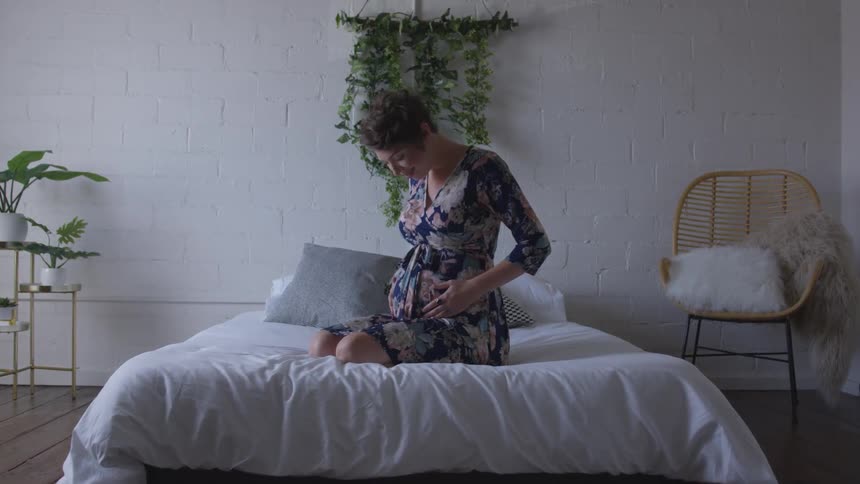 A pregnant woman kneels on a bed and feels her stomach smiling in anticipation
