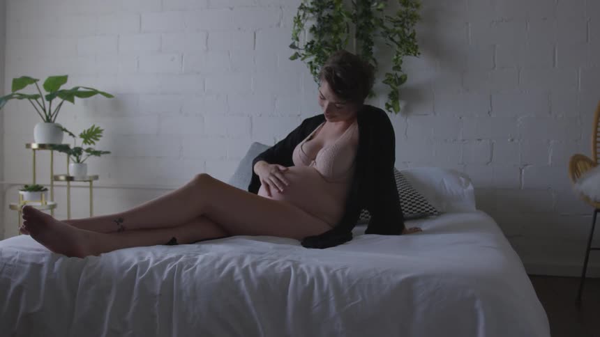 Pregnant woman sits on bed rubbing belly