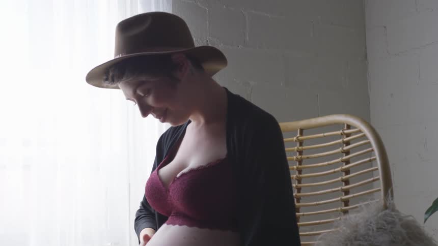 A pregnant woman wearing underwear and a hat rubs her stomach while smiling