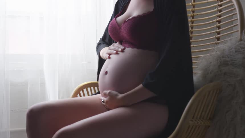 Pregnant woman wearing lingerie and a hat rubbing her stomach while sitting in a chair
