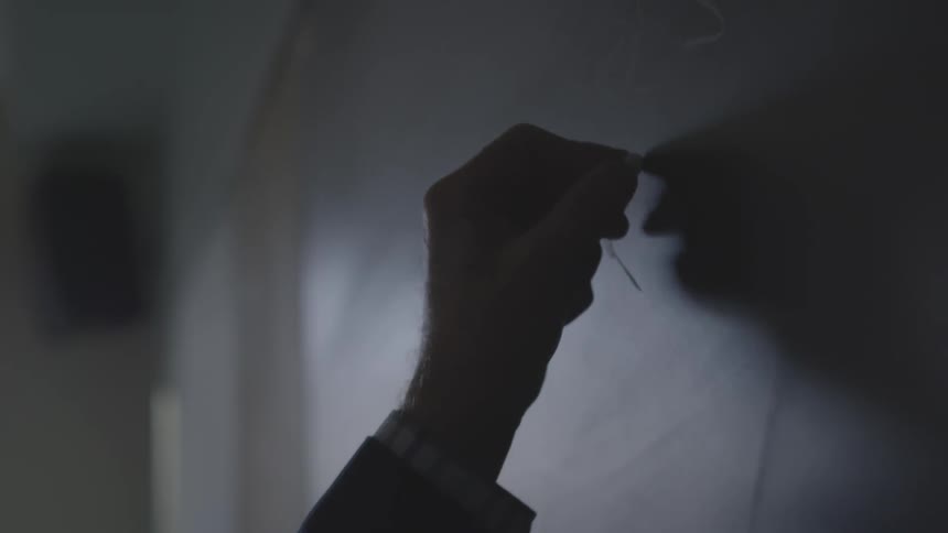 Person writes on chalkboard with chalk in dim room