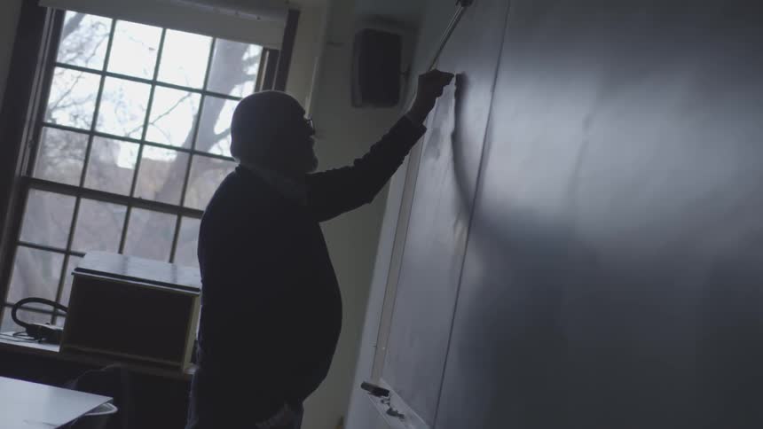 An old man writing on a chalkboard in a school