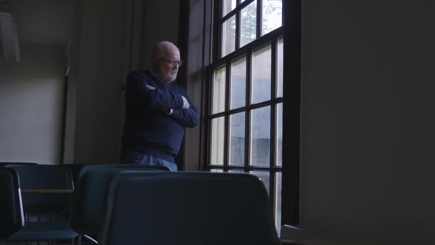 Elderly man standing with his arms crossed looking out a window