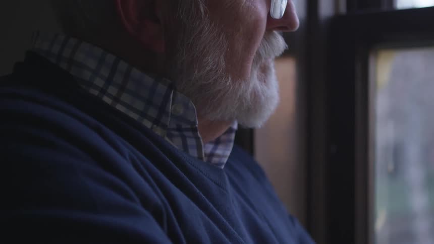 A bespectacled man with a white beard looks out of a window