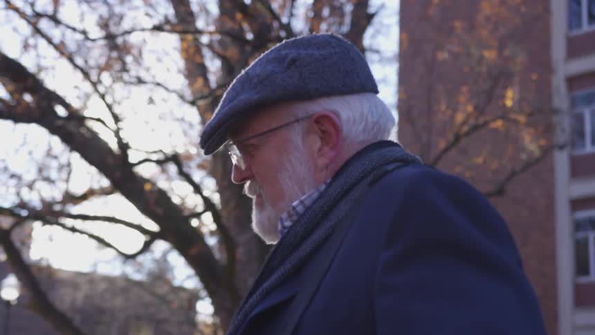 Old man passes by tree while sunlight shine on his face in city