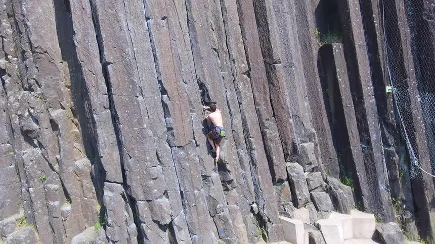 A solitary man without a shirt on is climbing up the rock face of of a tall mountainous wall