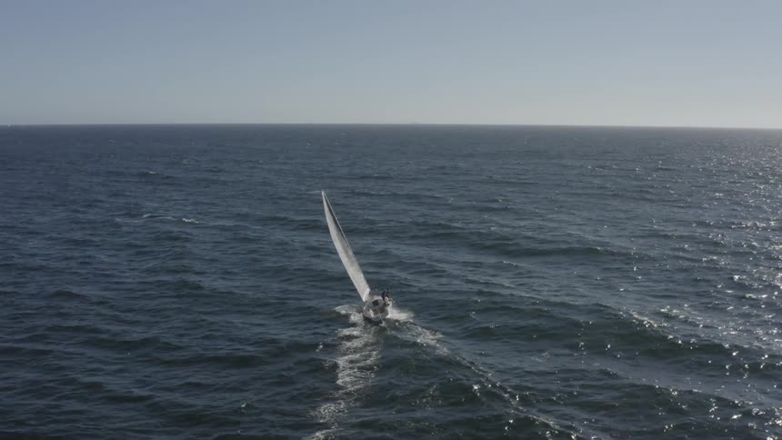 A sailboat swiftly cuts along the shallow waves in a wide open ocean space and two people sit at the edge of the bow of the boat