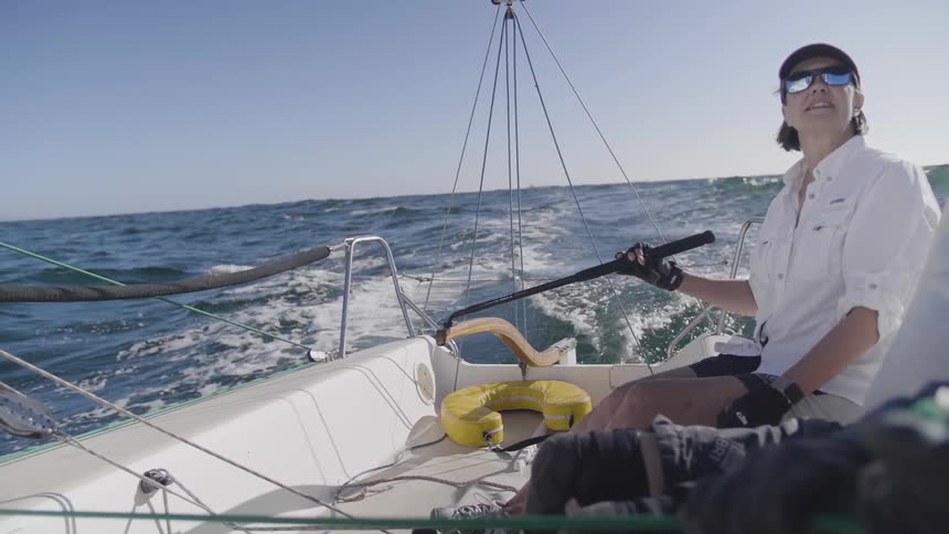 A person in a hat and sunglasses steers the rudder of a sailing boat