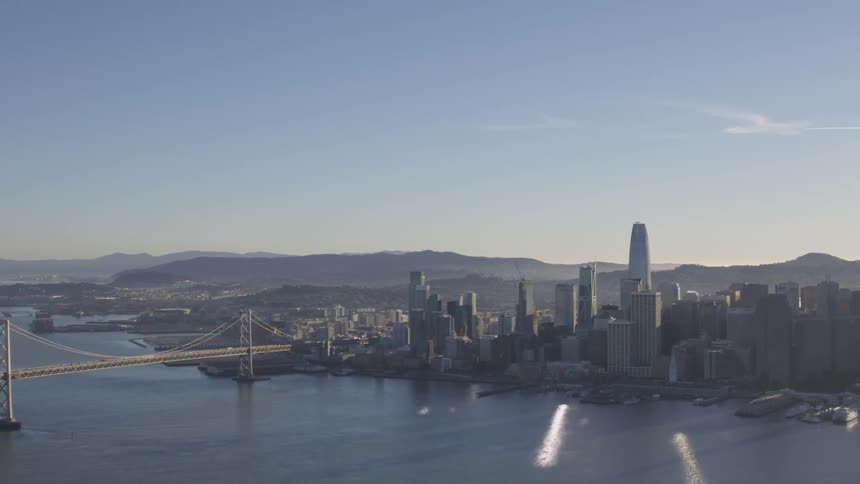 The sunshine reflects off the ocean harbor of a coastal city with skyscrapers and a bridge