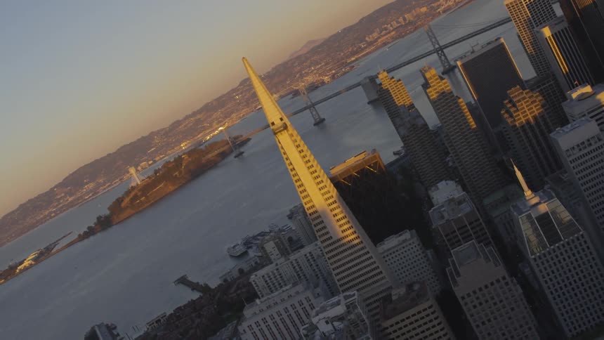 A city landscape set by a body of water is viewed from the air at low sun