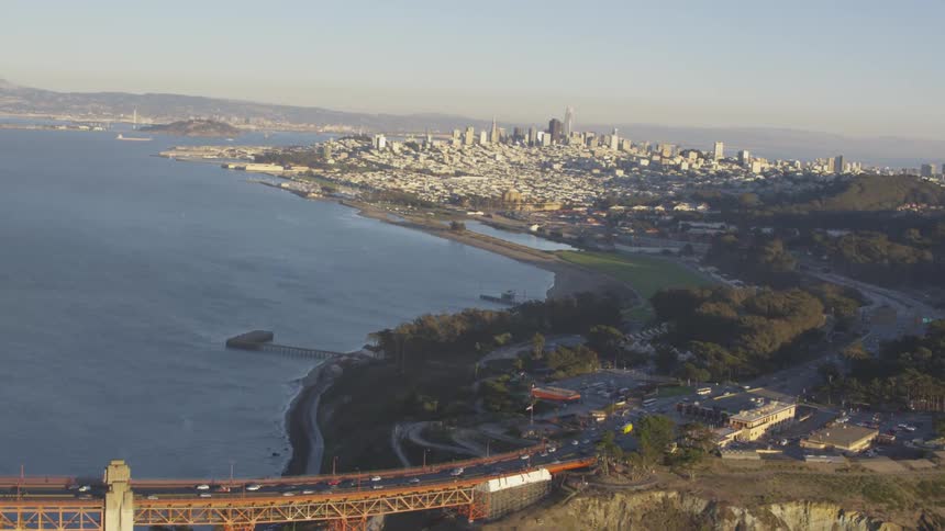 Cars drive on and off a bridge over the ocean with a city skyline in the background