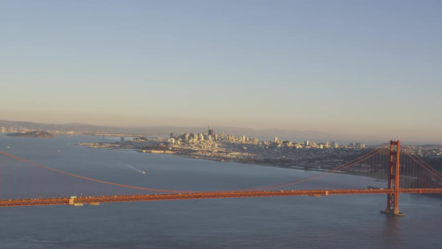 A suspension bridge crosses a wide river as traffic moves across it against the backdrop of a city skyline