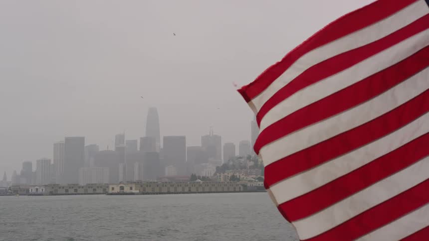 A flag waves over the water with a city in the distance