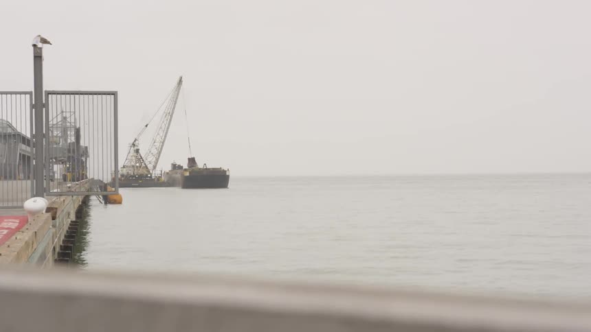 A crane on the waterfront loads a barge on a gray cloudy day