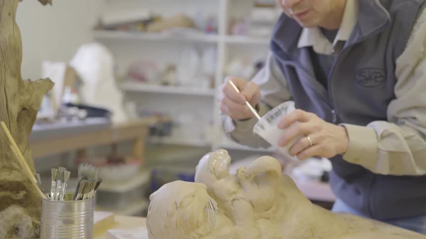 A man brushes a finish onto a clay sculpture of otters in his studio