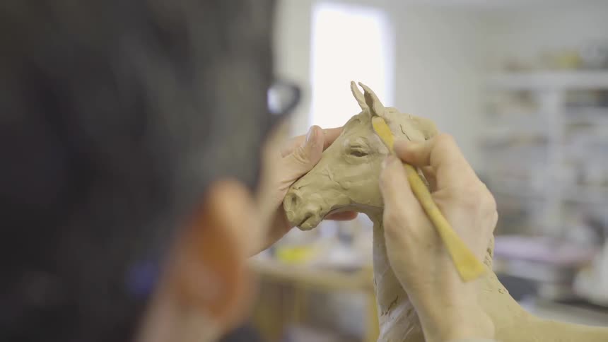 A woman is sculpting a horse while in a workshop during the day