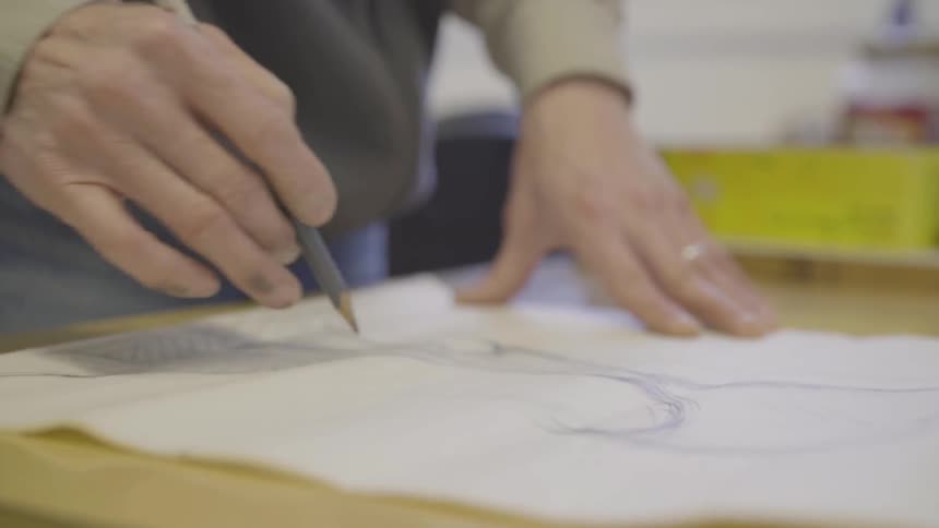 A man stands at a work table and uses a pencil to sketch on thin paper in a workshop