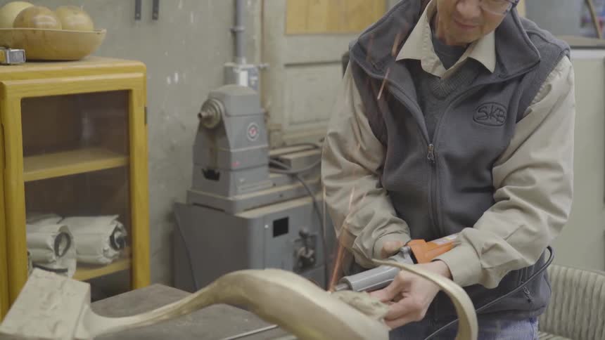 An older man in a workshop is slowly grinding away at an object which is causing sparks to wildly fly up into the air
