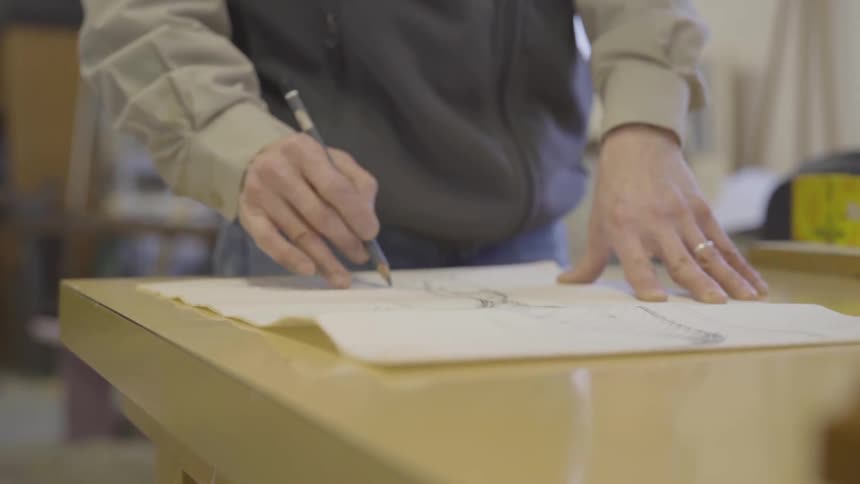 An artist stands at a table and sketches using a pencil on paper and smoothing edges with his thumb