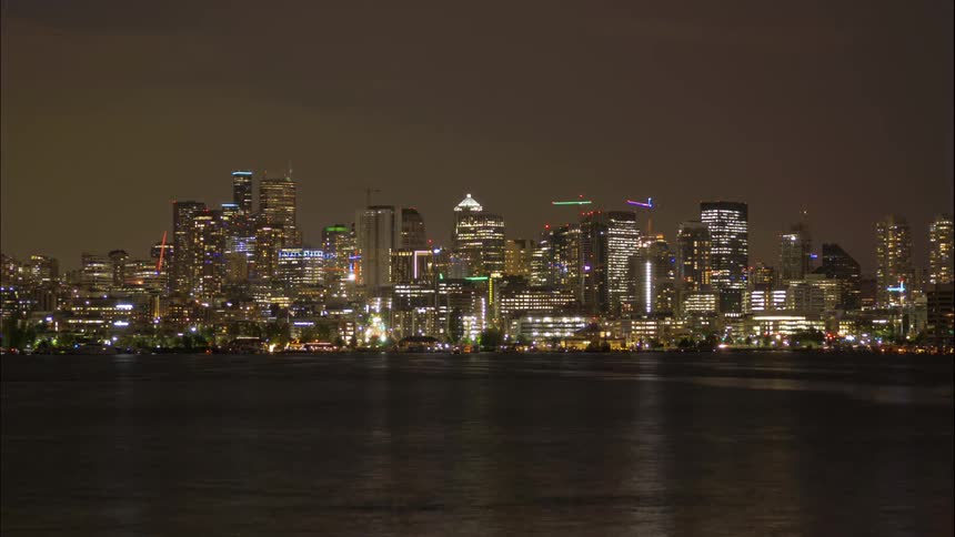 Boats float in a bay with city lights in the distance at night