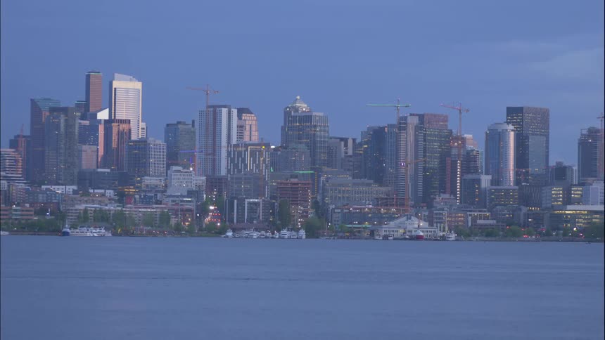 Boats pass through the harbor as lights from the city buildings and traffic twinkle at dusk