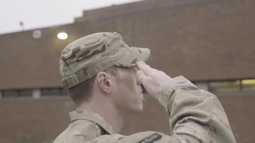 A white soldier in camouflage fatigues saluting while standing outdoors